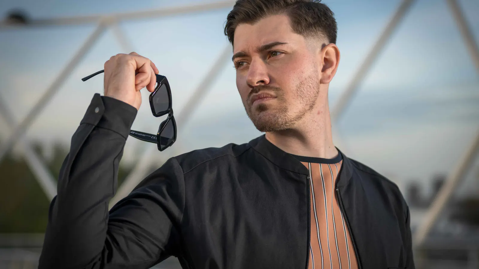 Junger Mann in schwarzer Jacke und gestreiftem Shirt hält eine Sonnenbrille in der Hand und blickt seitlich aus dem Bild. Modernes Fashion-Portrait vor urbaner Architektur.