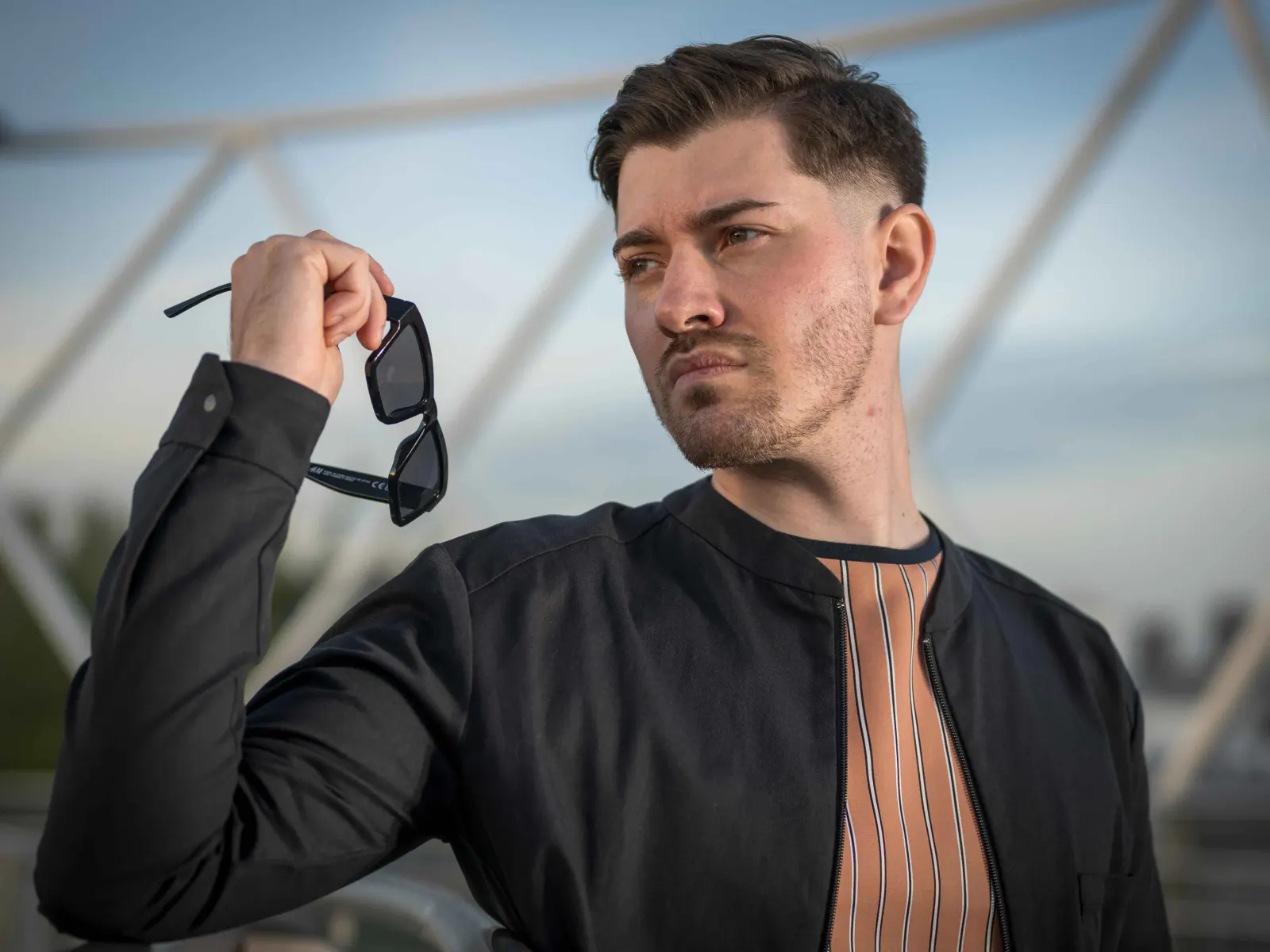 Junger Mann in schwarzer Jacke und gestreiftem Shirt hält eine Sonnenbrille in der Hand und blickt seitlich aus dem Bild. Modernes Fashion-Portrait vor urbaner Architektur.