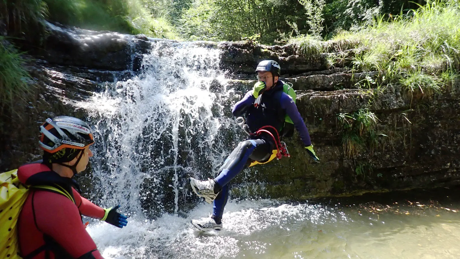 Eine Person im Flug bevor sie im Wasser landet mit einem kleinen Wasserfall im Hintergrund.