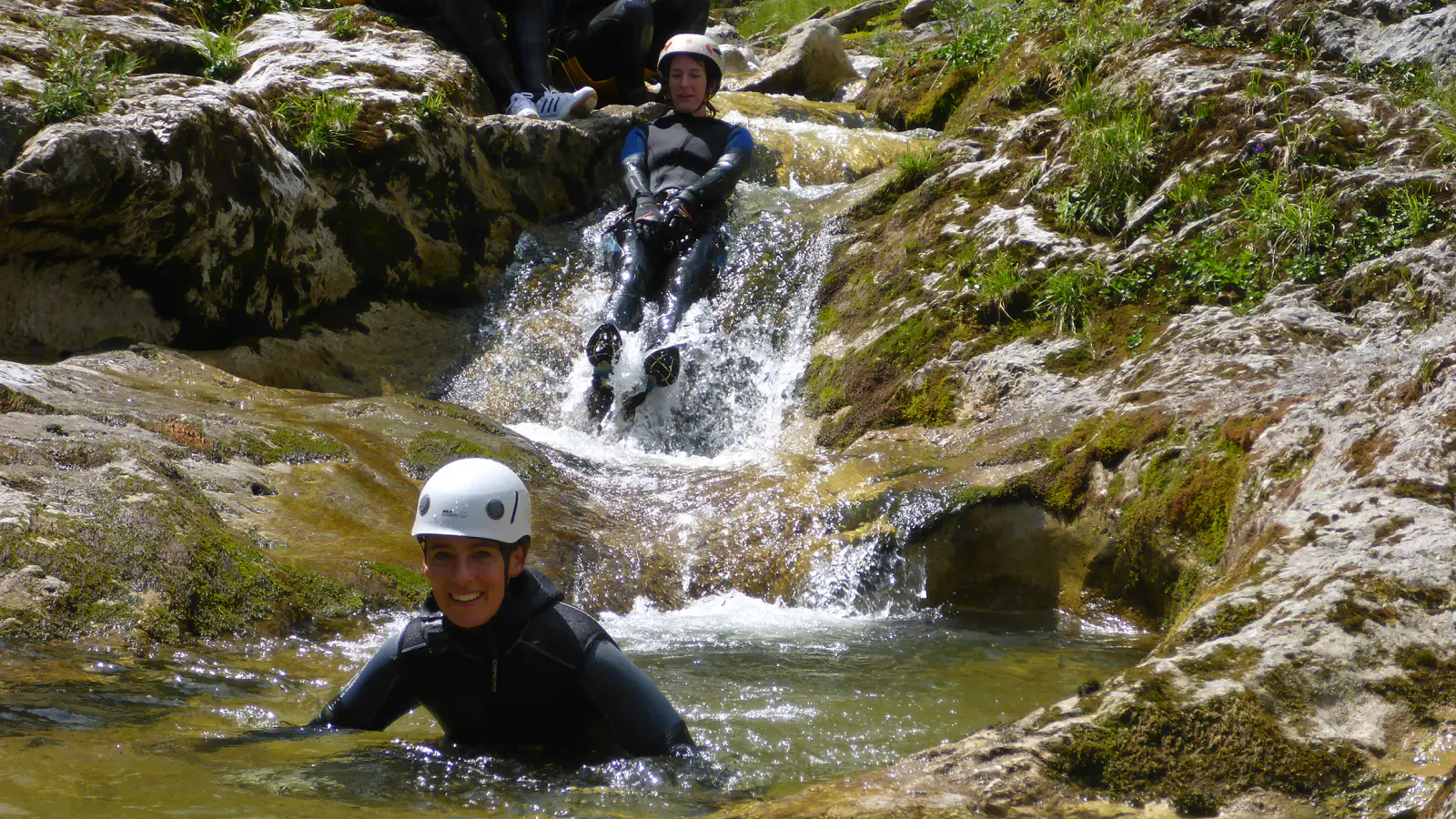 Eine Canyonautin sitzt im Wasser während eine andere hinter ihr einen Felsen hinunterrutscht