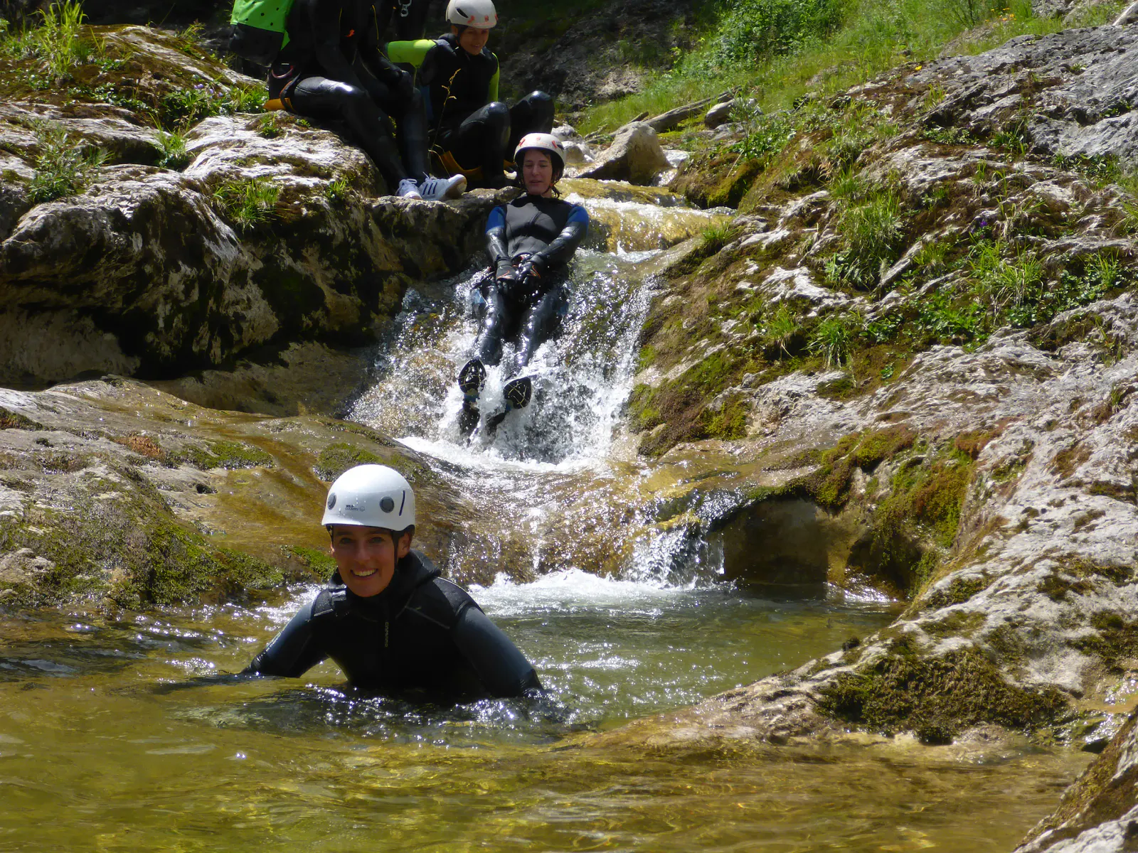 Eine Canyonautin sitzt im Wasser während eine andere hinter ihr einen Felsen hinunterrutscht