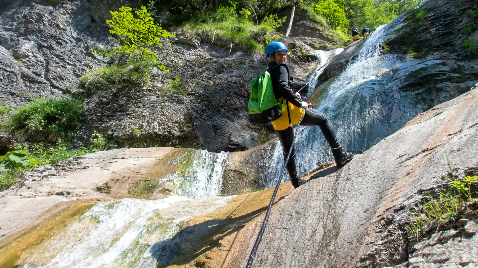Eine Person seilt sich neben einem Wasserfall ab und lächelt in die Kamera.