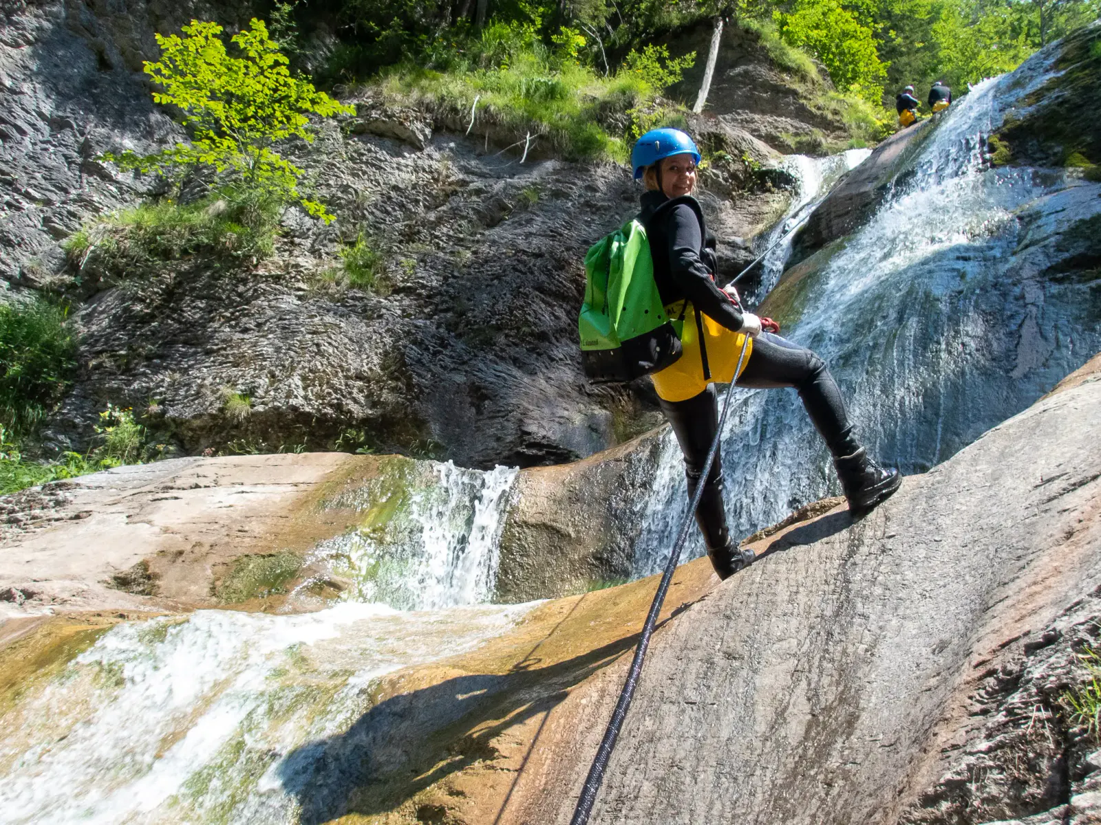 Eine Person seilt sich neben einem Wasserfall ab und lächelt in die Kamera.