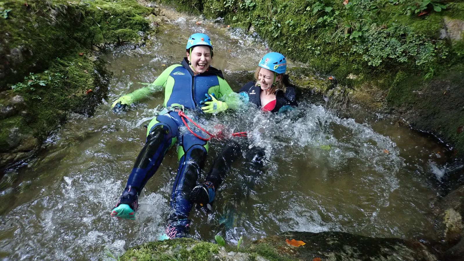 Zwei Personen in Canyoning-Ausrüstung liegen lachend im Gebirgsbach.