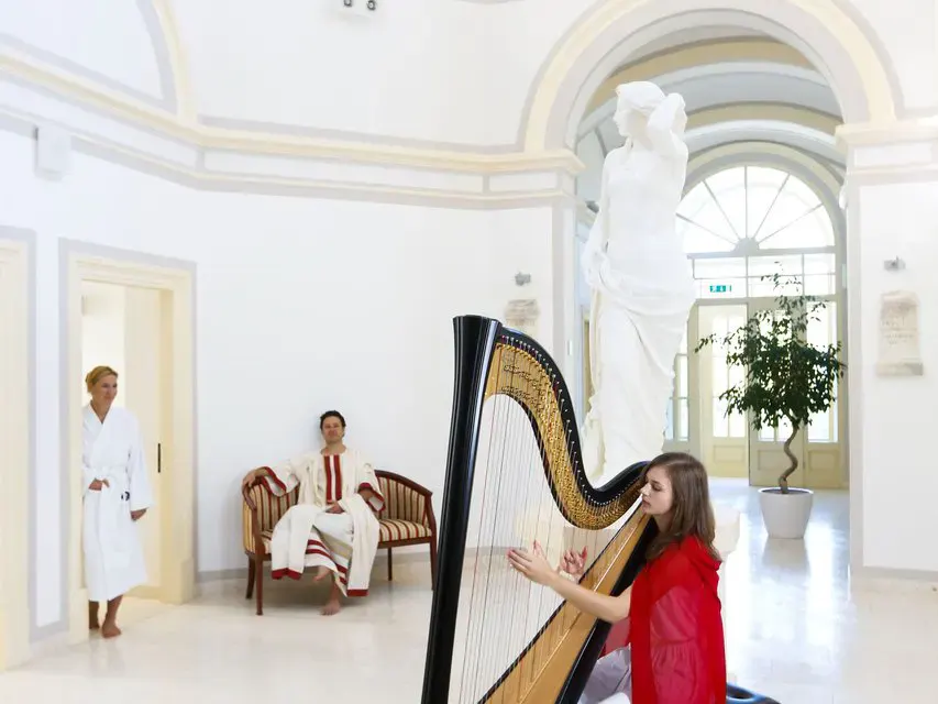 Eine Harfespielerin und zwei Gäste im Bademantel im historischen Teil des Wellnessbereiches im Rimske Terme Hotel Rimski dvor