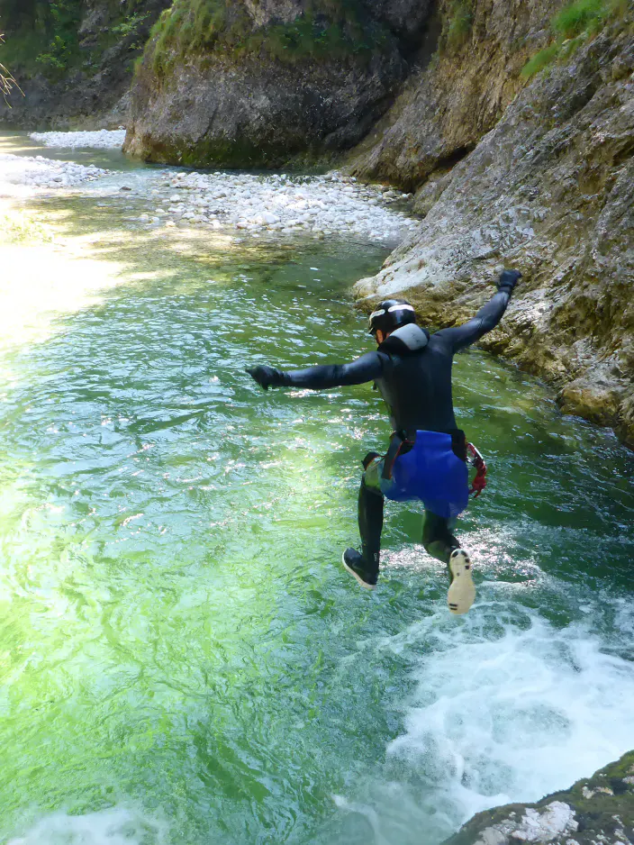 Eine Person in Neoprenanzug, Gurt und Helm springt in einer Schlucht von einem Felsen in türkises Wasser.