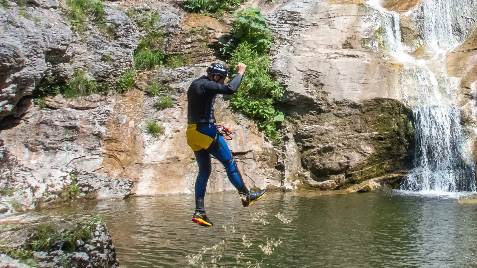 Eine Person während dem Sprung ins Wasser mit einem Wasserfall im Hintergrund.