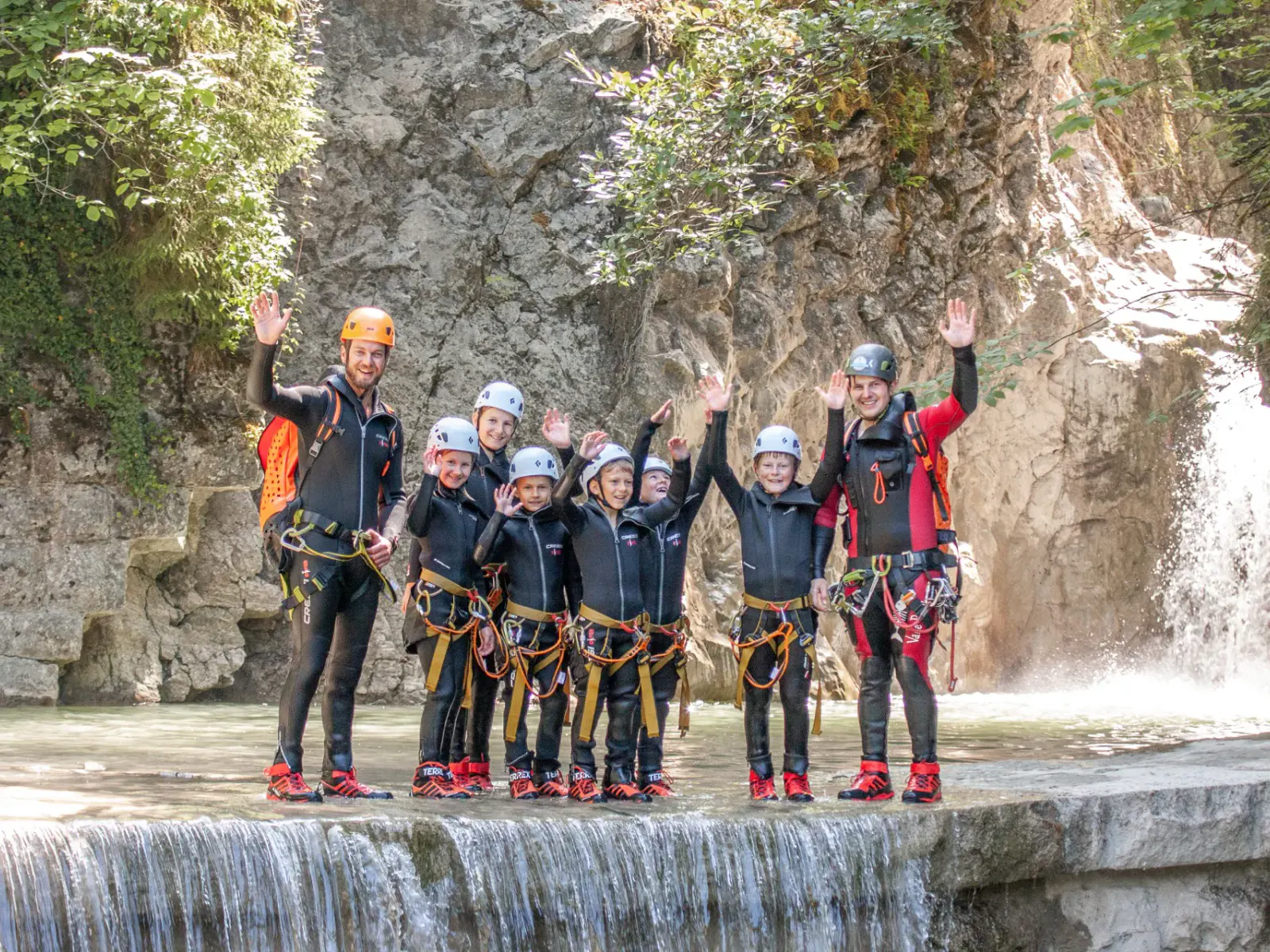 Kinder & Erwachsene stehen nebeneinander über einem kleine Wasserfall in Canyoningausrüstung