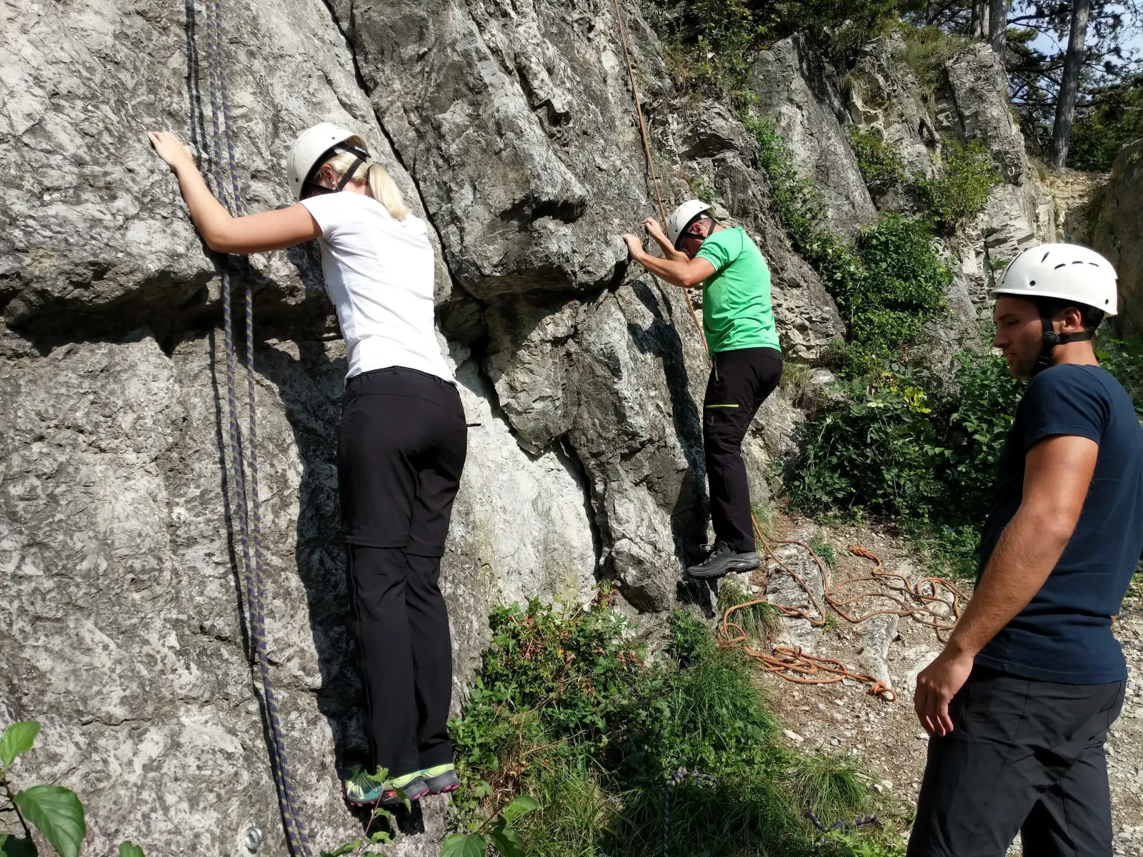 Zwei Personen bouldern an einer Felswand während eine dritte Person ihre Technik kontrolliert.