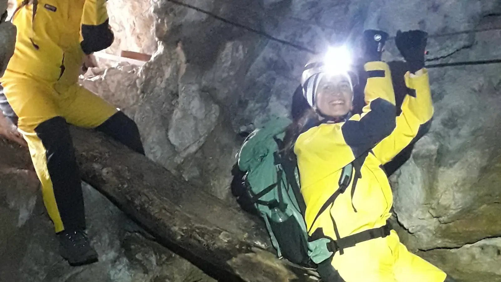 Zwei Personen in Höhlenanzügen rutschen über einen über eine Schlucht gelegten Baumstamm während sie sich an einem Seil über ihnen anhalten.