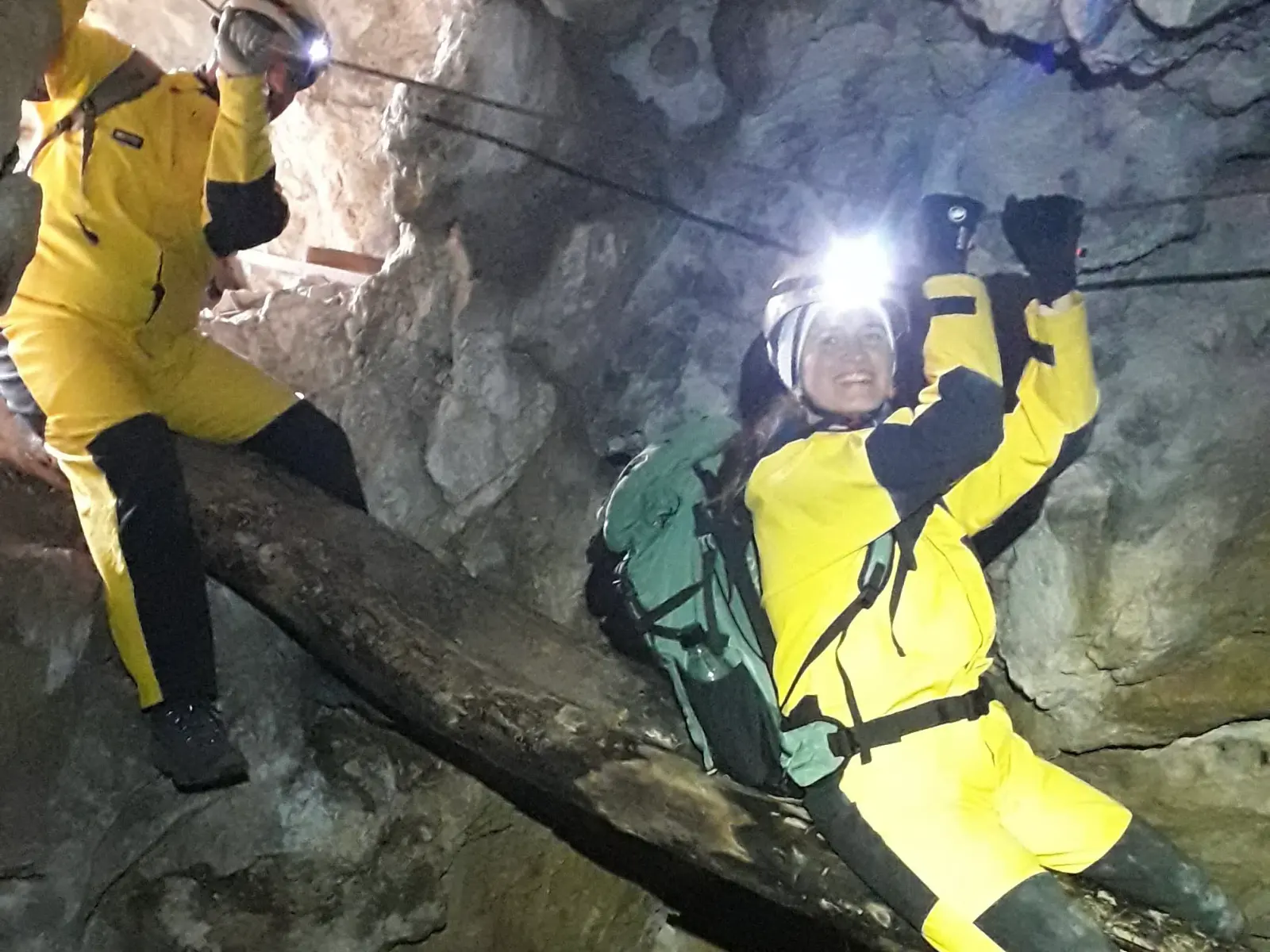 Zwei Personen in Höhlenanzügen rutschen über einen über eine Schlucht gelegten Baumstamm während sie sich an einem Seil über ihnen anhalten.