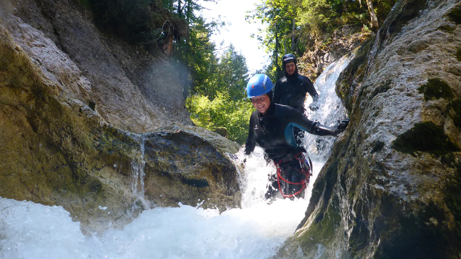Zwei Personen wandern in Neoprenanzügen durch eine Schlucht, in der das Wasser schäumt.