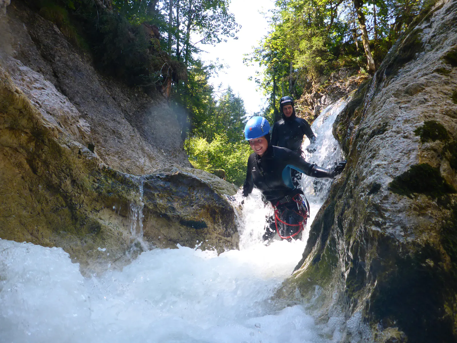 Zwei Personen wandern in Neoprenanzügen durch eine Schlucht, in der das Wasser schäumt.